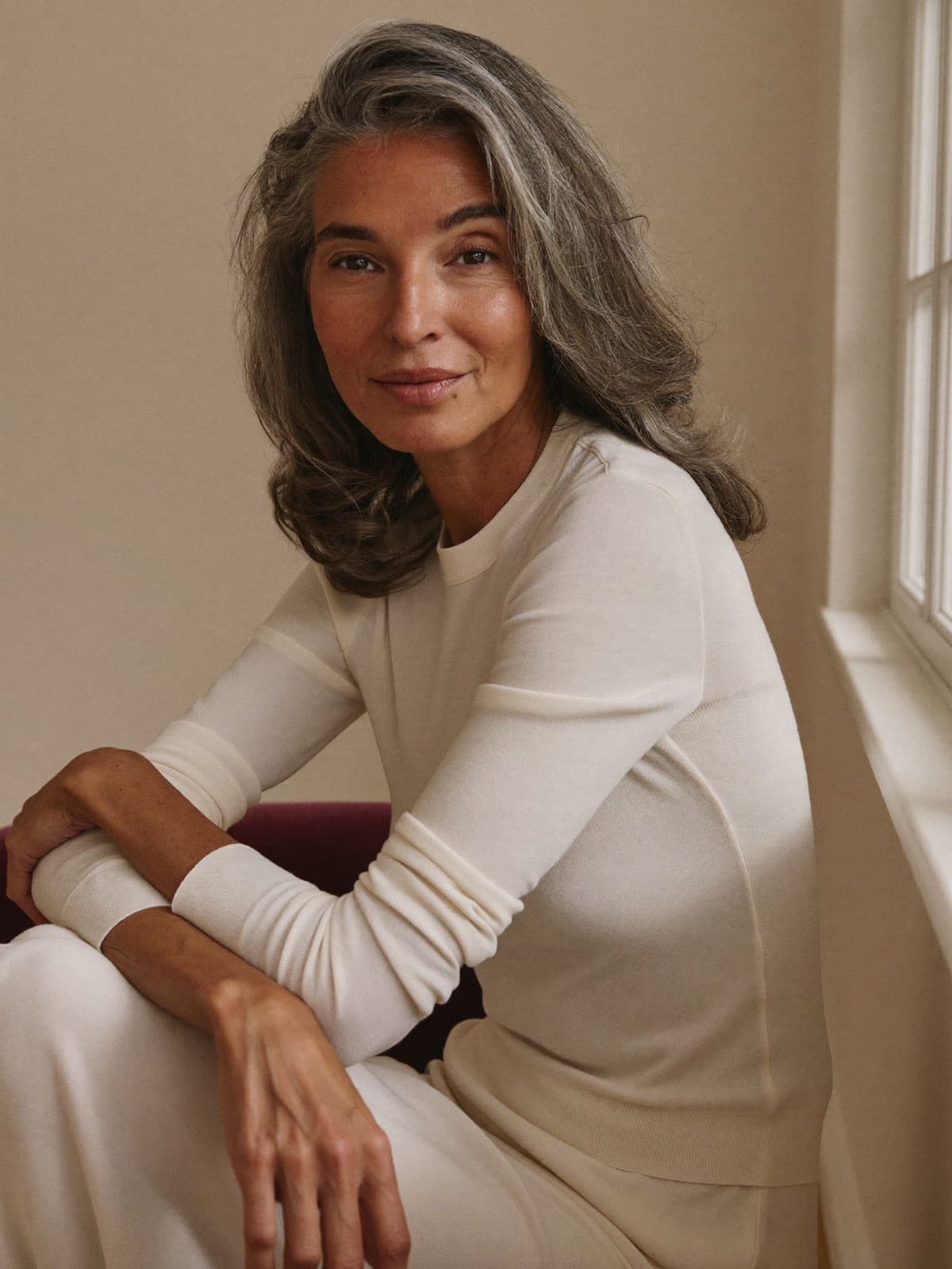 A woman in her fifties with silver hair, sitting by a window in soft daylight