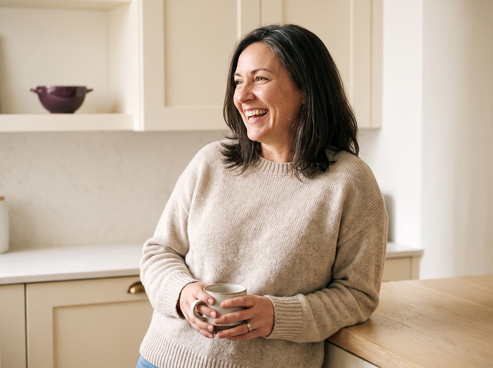 A woman in her late forties laughing over a cup of coffee in her kitchen